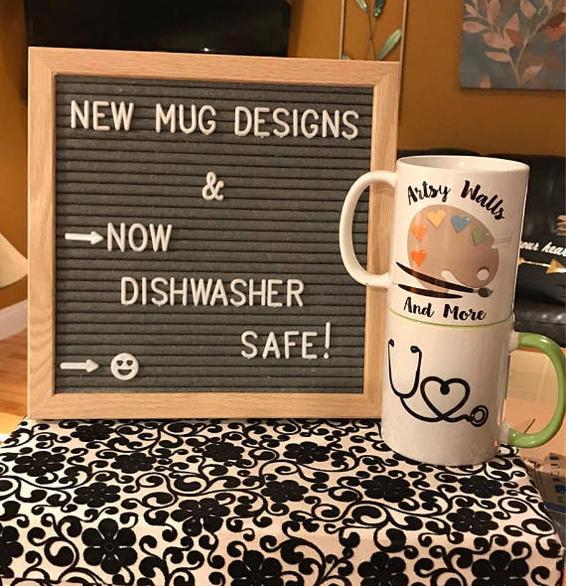 May include: A sign that reads "NEW MUG DESIGNS & NOW DISHWASHER SAFE!" with two mugs stacked on top of each other. The top mug has a painting palette design with the text "Artsy Walls" and the bottom mug has a stethoscope design with the text "And More".