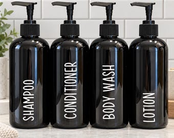 May include: Four black cylindrical plastic bottles with black pump dispensers. Each bottle features white vertical text labels reading SHAMPOO, CONDITIONER, BODY WASH, and LOTION. The bottles are arranged in a row on a light surface.