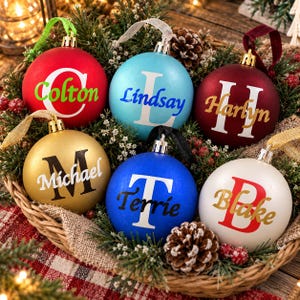 May include: Six round Christmas ornaments in red, light blue, dark red, gold, blue, and white. Each ornament features a large white or colored initial letter overlaid with a name in a contrasting script font, suspended by ribbons of various colors.