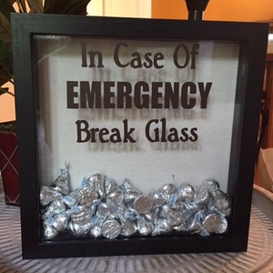 May include: A black shadow box with the text "In Case Of EMERGENCY Break Glass" and filled with silver foil-wrapped chocolate kisses.