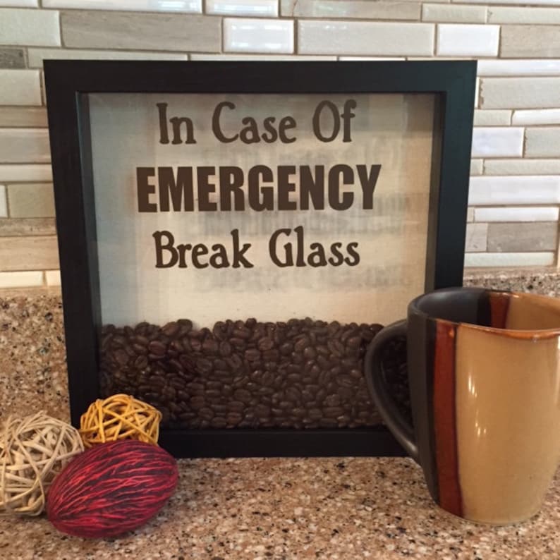 May include: A framed sign with the text "In Case Of EMERGENCY Break Glass" and a background of coffee beans. The sign is sitting on a counter next to a brown and tan coffee mug.
