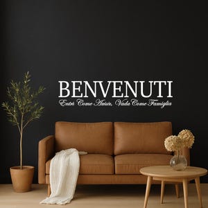 May include: A brown leather sofa with a white blanket draped over it, set against a dark wall. The wall features the text "BENVENUTI Entri Come Amici, Vada Come Famiglia" in white. A small wooden table and a potted plant are also in the scene.