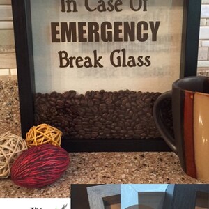 May include: A black framed shadow box with the text "In Case Of EMERGENCY Break Glass" and filled with coffee beans. The shadow box is sitting on a counter with a coffee mug and decorative balls.
