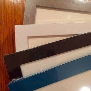 May include: Four picture frames stacked on top of each other. The top frame is a light gray, the second frame is white, the third frame is black, and the bottom frame is a dark teal blue.