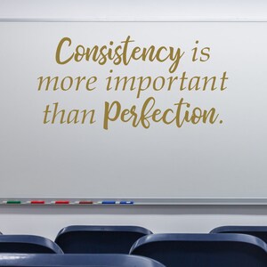 May include: Whiteboard with the quote "Consistency is more important than perfection." written in gold cursive lettering. The whiteboard is mounted on a wall above a row of blue chairs.
