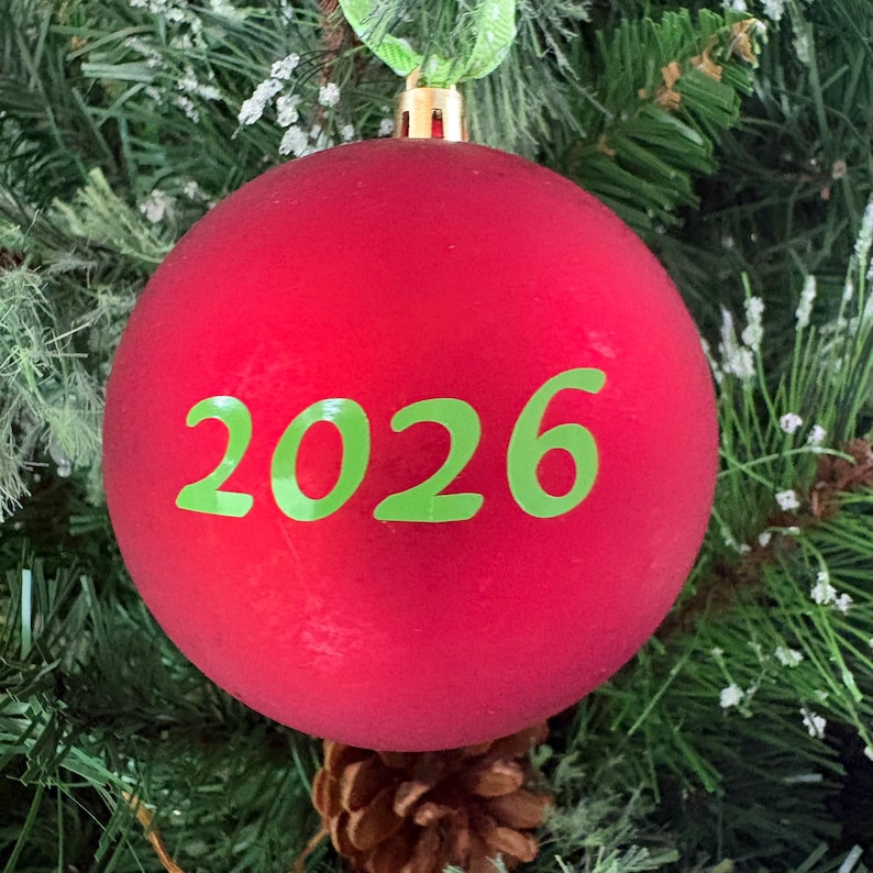 Może przedstawiać: Sferyczna czerwona bombka choinkowa z nadrukowanym na środku rokiem 2026 w jaskrawym limonkowym kolorze. Ozdoba posiada złotą metalową nasadkę u g&oacute;ry z zieloną wstążką do zawieszenia na gałęzi drzewa.