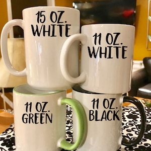 May include: Four ceramic mugs with different sizes and colors. The mugs are stacked on top of each other. The top mug is white and says "15 oz. WHITE". The second mug is white and says "11 oz. WHITE". The third mug is white with a green rim and says "11 oz. GREEN". The bottom mug is white with a black rim and says "11 oz. BLACK".