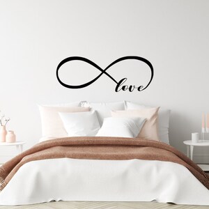 May include: A black infinity symbol with the word "love" written inside, displayed on a white wall above a bed with white bedding and a brown throw blanket.