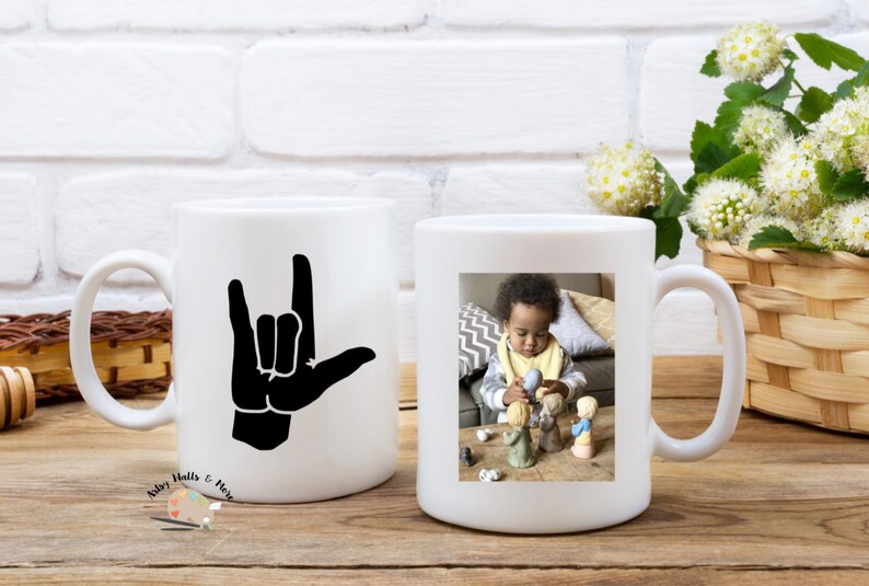 May include: Two white ceramic mugs with handles. One mug has a black silhouette of the American Sign Language symbol for "I love you". The other mug has a photo of a young child playing with toys.