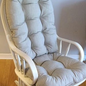 May include: A white wooden rocking chair with a light gray, tufted cushion. The chair has a high back and curved armrests. The seat and back cushions are made of a soft, textured fabric. The chair is designed for comfort and relaxation.
