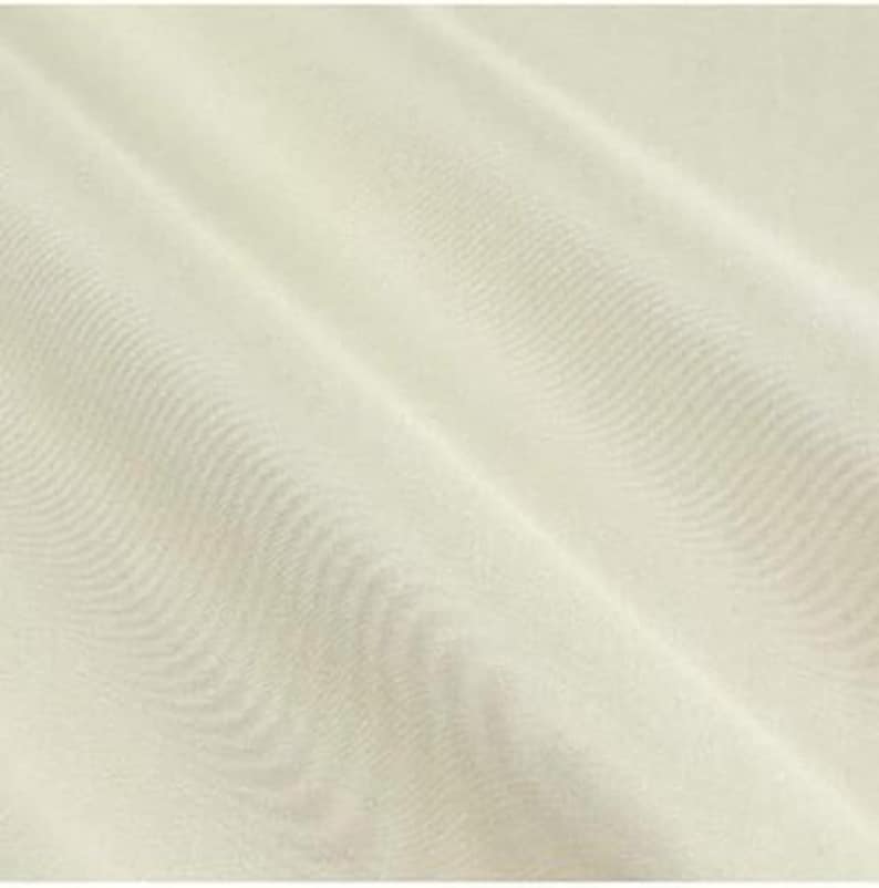 Op de afbeelding: Een effen gebroken witte stof met een zichtbare geweven textuur. Het materiaal vertoont diagonale plooien die zachte schaduwen en highlights over het oppervlak cre&euml;ren, wat het consistente weefpatroon en de neutrale, lichte cr&egrave;mekleur benadrukt.