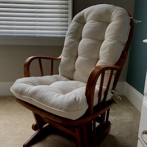 May include: A wooden rocking chair with a cream-colored, tufted cushion. The chair has a curved back and armrests, and the wooden frame has a warm, natural finish. The chair is designed for comfort and relaxation.