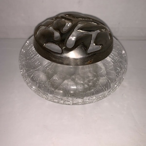 Puede incluir: Un tarro de vidrio transparente con un acabado craquelado y una tapa de metal plateado con un diseño floral decorativo. El tarro es perfecto para guardar potpourri u otros artículos decorativos.