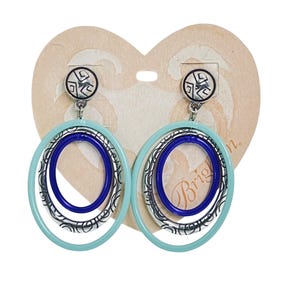 Puede incluir: Pendientes de forma ovalada con un anillo exterior azul claro, un anillo central de filigrana plateada y un anillo interior azul oscuro. Los pendientes tienen una peque&ntilde;a parte superior redonda con un dise&ntilde;o negro y plateado. Se muestran en una etiqueta en forma de coraz&oacute;n.