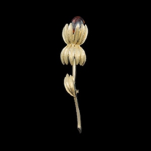 May include: A gold-toned flower brooch with a brown glass center. The flower has a long stem and a single leaf.