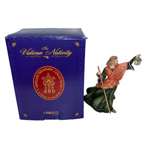 Puede incluir: Figura de cerámica de una persona con una túnica fluida, sosteniendo una linterna y un bastón, junto a una caja azul. La caja dice "The Vatican Nativity" y "Vatican Treasury Collection". La figura es verde, roja y dorada.
