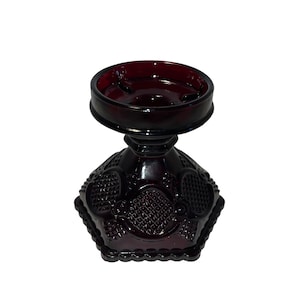 Puede incluir: Un portavelas de vidrio rojo oscuro con una base hexagonal y una parte superior redonda. La base tiene un patrón decorativo de corazones y espirales.