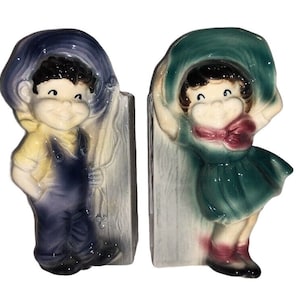 Puede incluir: Un par de figuritas de maceta de pared de cerámica vintage. Las figuritas son de un niño y una niña, ambos con sombreros. El niño lleva un atuendo azul y amarillo y la niña lleva un vestido verde con un lazo rojo. Las figuritas están unidas a un fondo blanco.