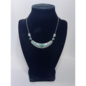 May include: Silver-toned necklace with turquoise-colored stones. The necklace features a curved centerpiece with a large oval stone in the center and smaller stones around it. The necklace is displayed on a black mannequin.