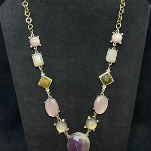 May include: A gold chain necklace with a variety of gemstone pendants, including pink, white, brown, and purple stones. The necklace features a large, round, purple stone pendant in the center.