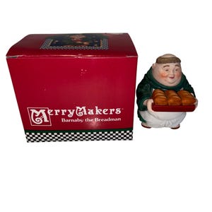 May include: A ceramic figurine of a jolly monk holding a tray of bread, next to a red box with the text "Merry Makers Barnaby the Breadman". The figurine is wearing a green robe and white apron. The box has a black and white checkered pattern.