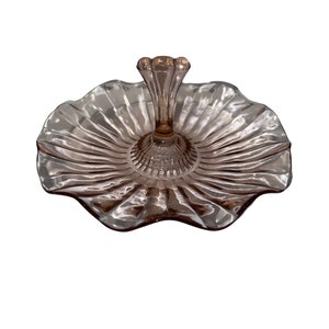 May include: A pink glass serving dish with a fluted edge and a central handle. The dish has a scalloped rim and a delicate, feminine design.