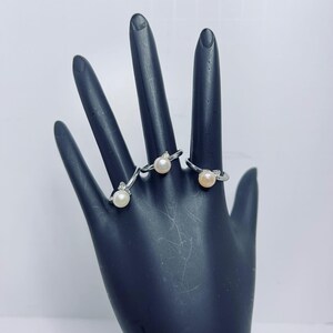 May include: Three silver rings, each featuring a single pearl and small accent stones, are displayed on a black hand-shaped display. The pearls have a creamy, off-white color.