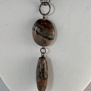 May include: A silver chain necklace with a brown and black marbled stone pendant. The pendant is a round disc with a smaller, cylindrical stone hanging from it.