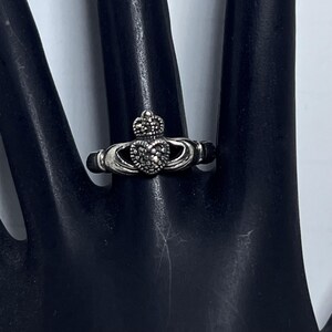 May include: A silver Claddagh ring featuring a central heart topped with a crown, flanked by two hands. The heart and crown are embellished with small, dark, textured stones, creating a detailed, metallic finish on the band and design elements.