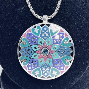 May include: A silver pendant necklace with a large, circular pendant. The pendant has a colourful, intricate design with teal, pink, and purple enamel, and a small blue gemstone in the centre. The necklace has a silver chain.