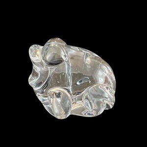 May include: A clear glass sculpture of a frog. The frog is sitting on its belly with its legs tucked underneath it. The sculpture is made of clear glass and has a smooth, polished finish.