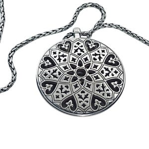 May include: A silver pendant necklace with a detailed, symmetrical design. The circular pendant features a pattern of hearts, floral elements, and geometric shapes. The chain is a woven style, complementing the pendant's ornate design.