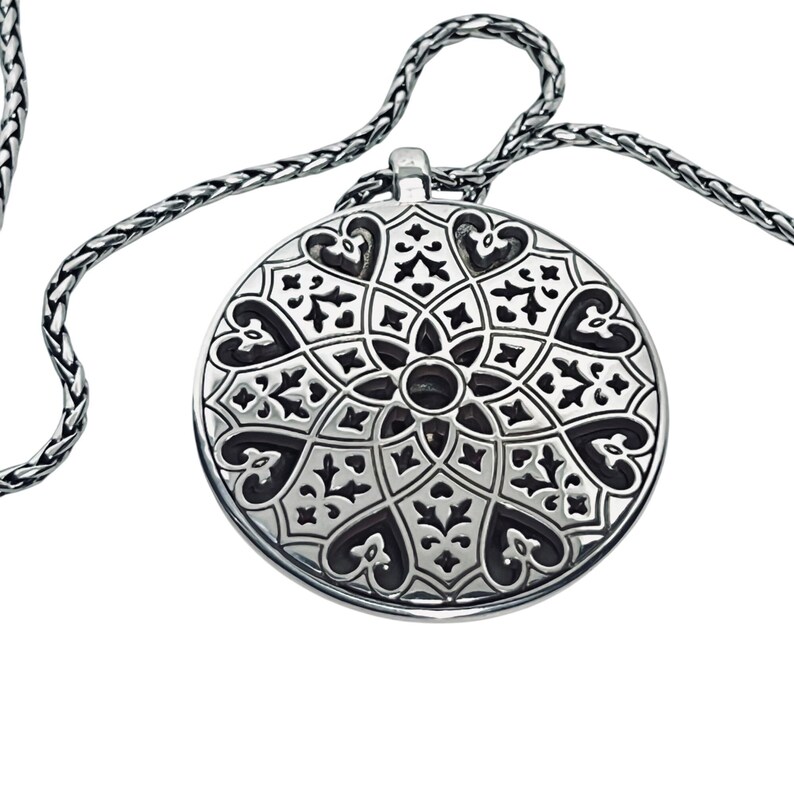 May include: A silver pendant necklace with a detailed, symmetrical design. The circular pendant features a pattern of hearts, floral elements, and geometric shapes. The chain is a woven style, complementing the pendant's ornate design.