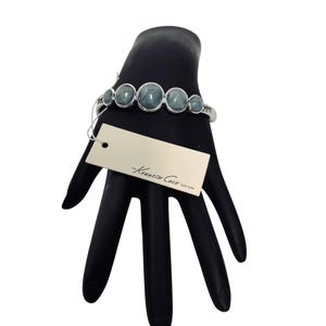 May include: Silver-tone bracelet with five oval, blue-green stones. The bracelet has a tag that reads "-Kenneth Cole NEW YORK". The bracelet is displayed on a black hand.