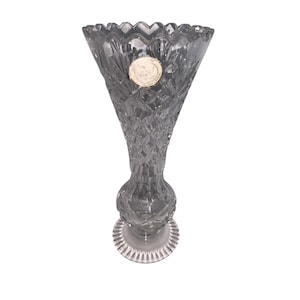 Puede incluir: Un jarrón de cristal transparente teñido de gris, con la parte superior acampanada y un cuerpo con diseño de diamantes. El jarrón tiene una base redonda y un borde decorativo. Una pequeña pegatina redonda está adherida a la parte delantera del jarrón.