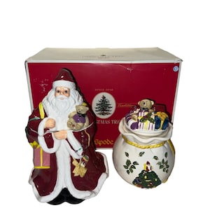 Op de afbeelding: Een vintage kerstman van keramiek van Christmas Tree Shops en een koekjespot in de vorm van een santasack. De kerstman houdt een teddybeer en een rol vast. De koekjespot is versierd met een kerstboom en een teddybeer. De doos is rood met de woorden "Christmas Tree Shops" en "Since 1918" erop gedrukt.