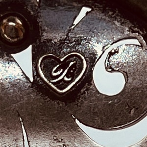 以下が含まれることがあります： ハート型のシルバーチャームのクローズアップ。チャームの中には「G」の文字が入っています。チャームはジュエリーに付いています。