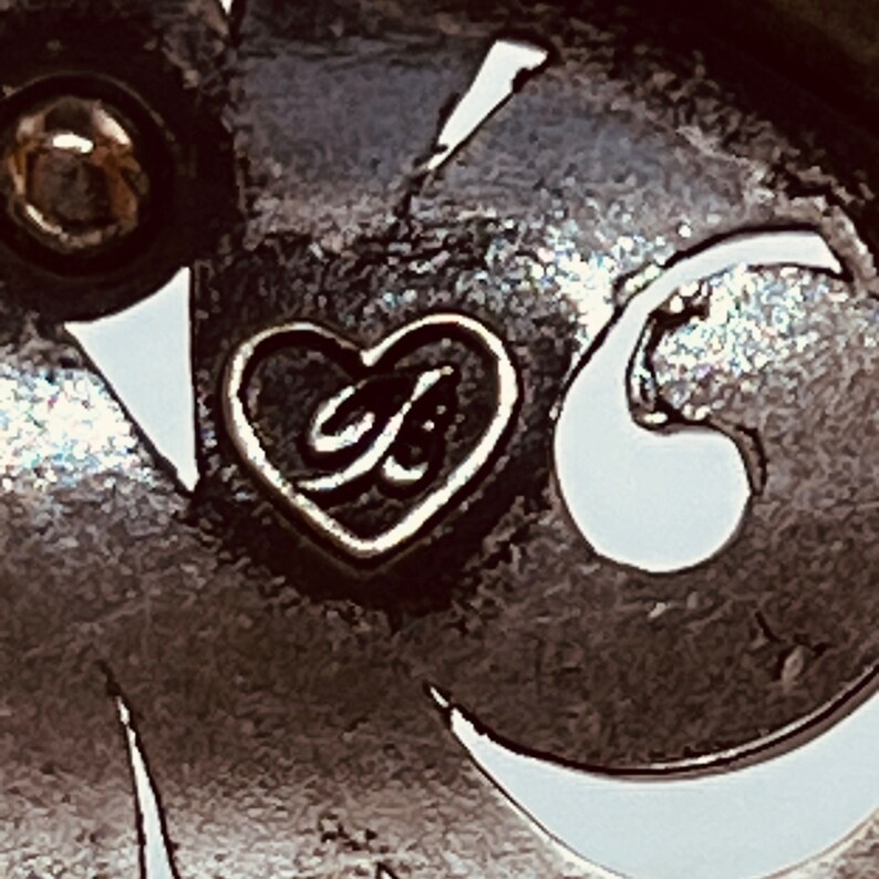 以下が含まれることがあります： ハート型のシルバーチャームのクローズアップ。チャームの中には「G」の文字が入っています。チャームはジュエリーに付いています。