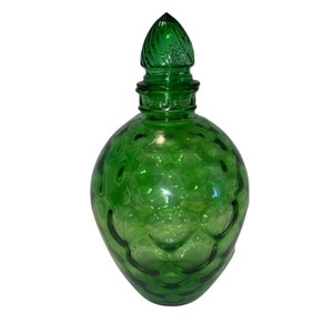 Puede incluir: Una botella de vidrio verde con una superficie texturizada y un tapón decorativo. La botella tiene un cuerpo redondo y un cuello alto y estrecho.