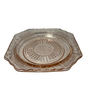 May include: A square, pink glass plate with a scalloped edge and a floral design in the center. The plate has a raised, sunburst pattern around the floral design.