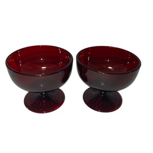 Può includere: Due coppette da dessert in vetro rosso rubino. Ogni coppetta ha una parte superiore arrotondata e aperta e poggia su un gambo corto con un motivo decorativo a forma di ventaglio. Le coppette sono in vetro colorato trasparente.