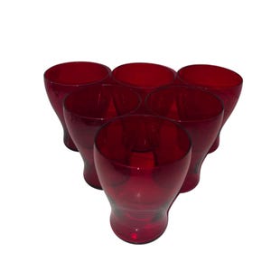 May include: Six ruby red drinking glasses arranged in a triangular shape. The glasses have a rounded base and a slightly flared rim. The color is a deep, transparent red, and the glasses appear to be made of glass.