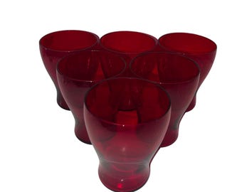 May include: Six ruby red drinking glasses arranged in a triangular shape. The glasses have a rounded base and a slightly flared rim. The color is a deep, transparent red, and the glasses appear to be made of glass.