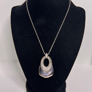 May include: Silver necklace with a pendant. The pendant has a textured, teardrop-shaped base and a sparkling, oval-shaped top. The necklace is displayed on a black mannequin, highlighting the jewellery's design and details.
