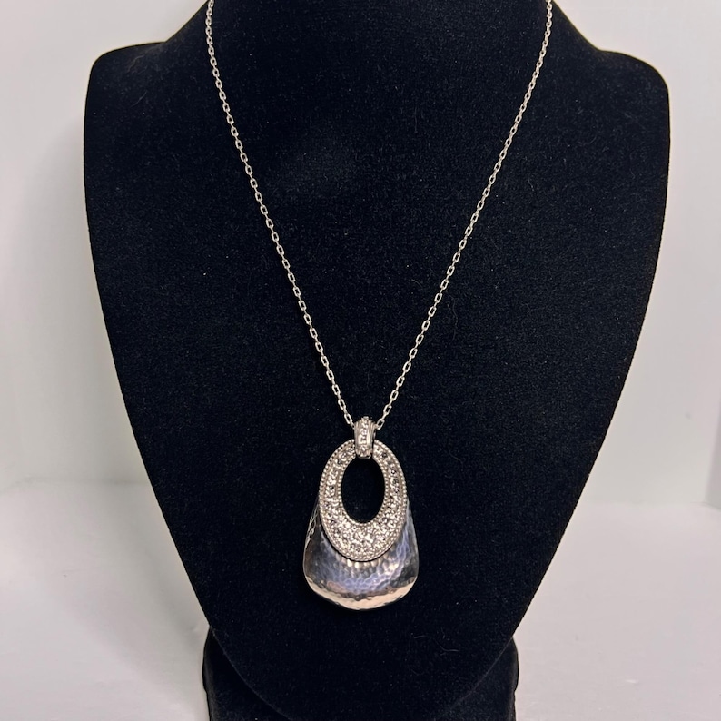 May include: Silver necklace with a pendant. The pendant has a textured, teardrop-shaped base and a sparkling, oval-shaped top. The necklace is displayed on a black mannequin, highlighting the jewellery's design and details.