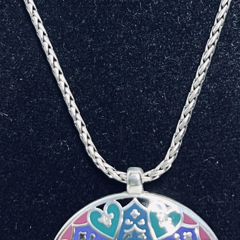 May include: Silver necklace with a detailed pendant. The pendant has a colourful, intricate design with pink, blue, and green accents. The chain is a woven style, and the pendant is suspended from a small loop.