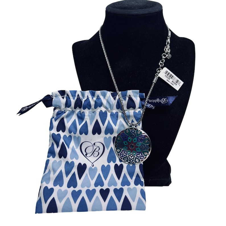 May include: A silver necklace with a large, circular pendant showcasing a colourful floral pattern. The necklace is displayed on a black mannequin, beside a blue drawstring bag with a heart design. A tag is attached to the necklace.