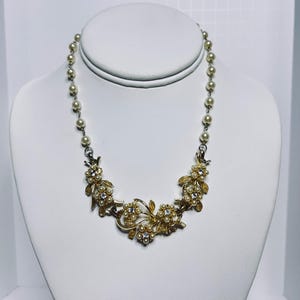 May include: A vintage necklace with faux pearls and gold-tone floral accents. The necklace features a strand of small, round pearls connected to a decorative centerpiece with gold-colored flowers, clear rhinestones, and more pearls. The necklace is displayed on a white mannequin.