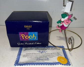 May include: A collectible Piglet ornament from the Grolier President's Edition, featuring a pink Piglet character with a green hat and mittens. The ornament is displayed on a gold-colored stand, with a blue box and a certificate.