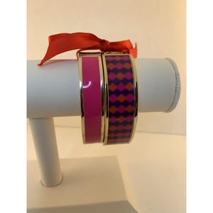 May include: Two gold-toned bangles displayed on a white stand, one solid magenta, the other patterned with orange, purple, and magenta diamonds. A red ribbon is tied at the top of the stand.